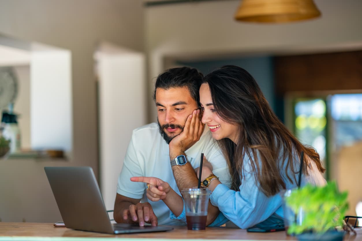 A couple smiles while looking at a laptop in a cozy living room. The woman points at the screen, with an iced coffee on the table, conveying a relaxed mood.