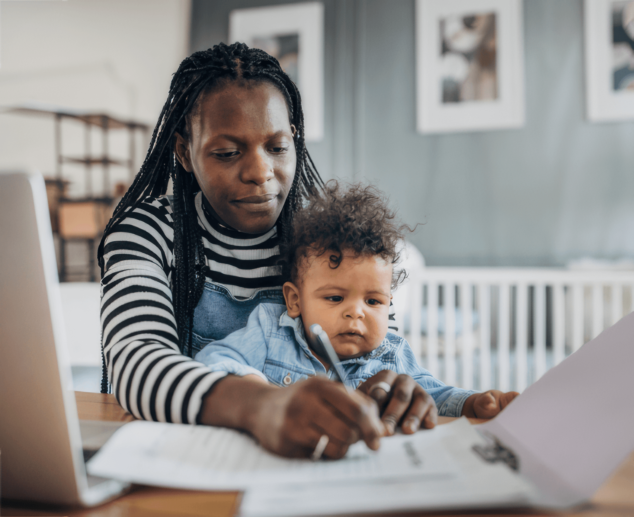 A woman with braided hair focuses on paperwork at a table, holding a pen, with a toddler on her lap. The setting is a cozy, bright home.