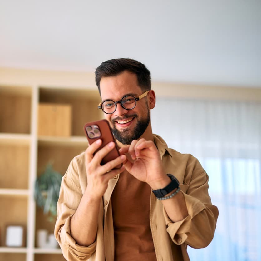 A bearded man wearing tortoiseshell glasses and a tan shirt smiles while using his smartphone in a bright, modern living space. Built-in shelving with plants and books creates a warm, minimalist backdrop.