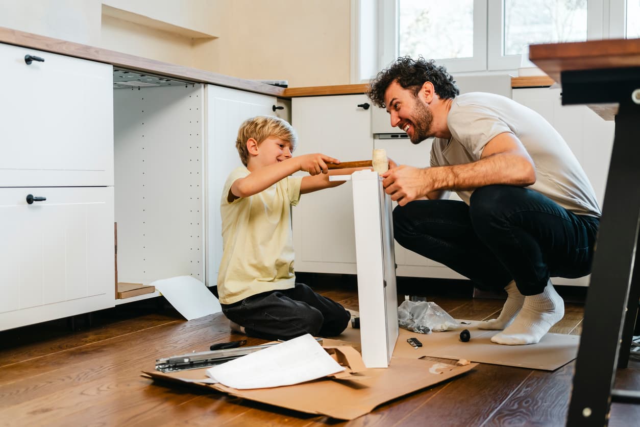 Father and son assembling furniture in a bright kitchen. The boy uses a mallet while the father assists, both smiling and enjoying their teamwork.